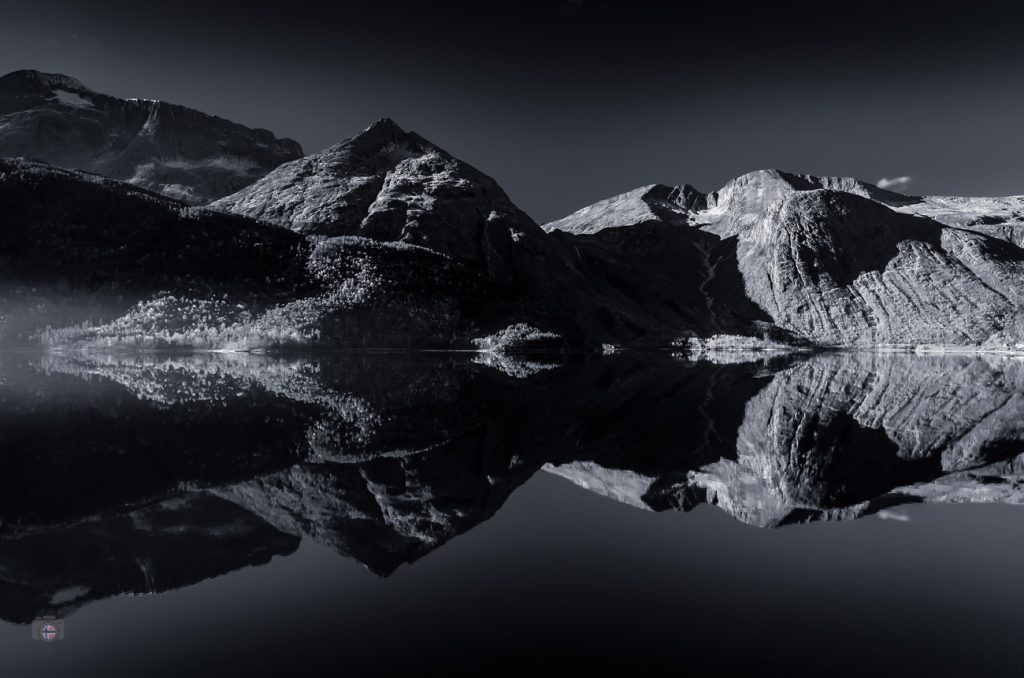 Schwarzweiß-Foto eines stillen Bergsees in Norwegen am frühen Morgen mit klarer Spiegelung der umliegenden Berge und leichtem Nebel über dem Wasser.