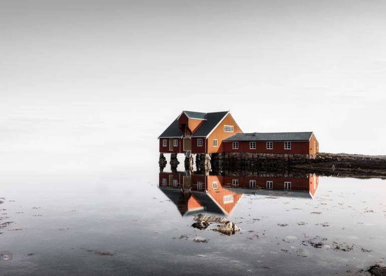 Traditionelles rotbraunes Fischerhaus an einer ruhigen Wasserfläche mit nahezu symmetrischer Spiegelung.