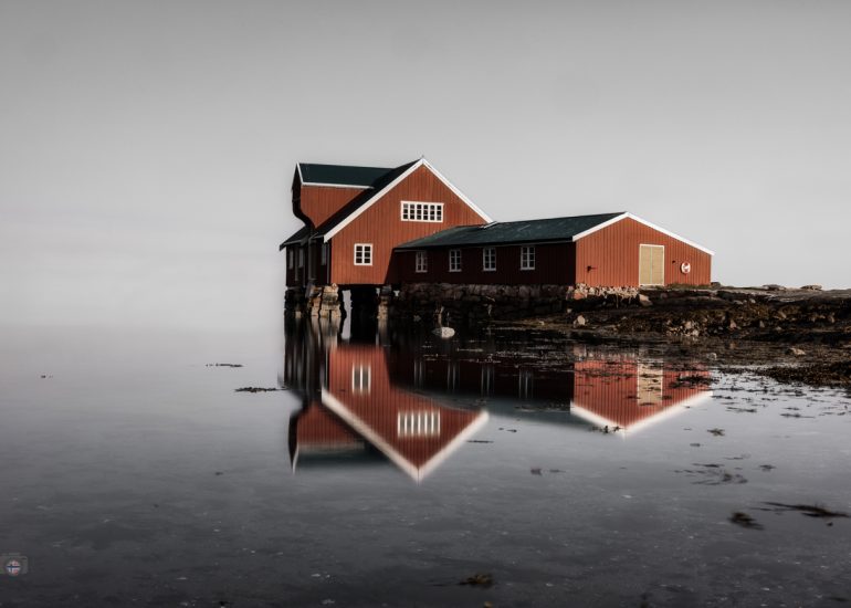 Traditionelles rotbraunes Fischerhaus an einer ruhigen Wasserfläche mit nahezu symmetrischer Spiegelung.