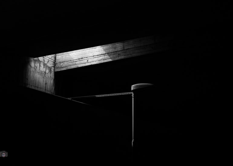 Schwarzweißfoto unter einer Brücke in Oslo. Ein Sonnenstrahl beleuchtet Beton und eine Lampe, der Hintergrund liegt vollständig im Schatten.
