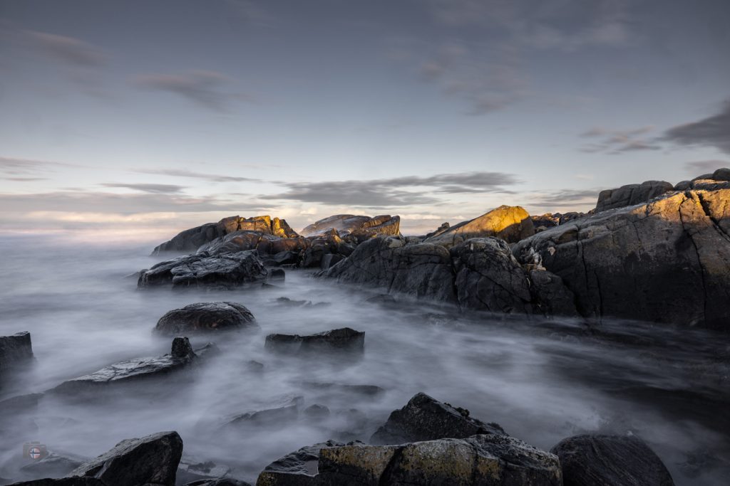 Steine im Meer bei Sonnenaufgang. Weiches Winterlicht und geglättetes Wasser durch Langzeitbelichtung erzeugen eine ruhige, reduzierte Stimmung zwischen Bewegung und Stille.