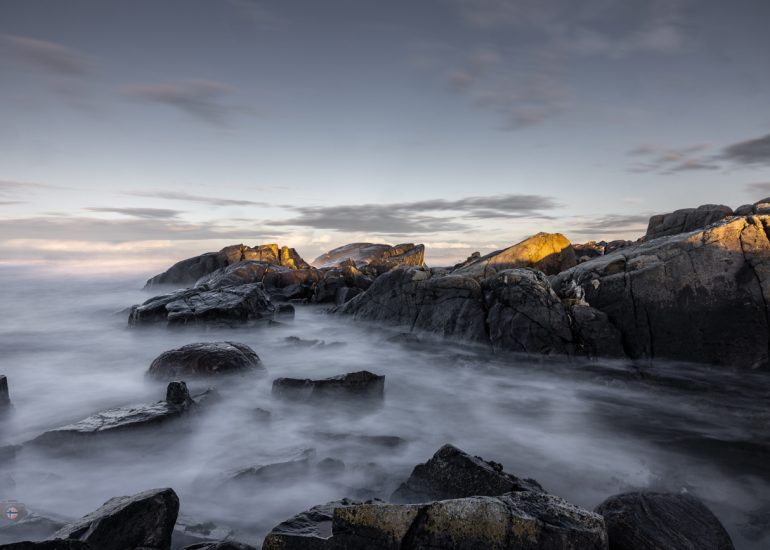 Steine im Meer bei Sonnenaufgang. Weiches Winterlicht und geglättetes Wasser durch Langzeitbelichtung erzeugen eine ruhige, reduzierte Stimmung zwischen Bewegung und Stille.
