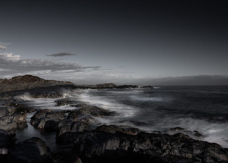 Dunkle Felsen an einer norwegischen Küste mit weich gezeichnetem Meerwasser unter einem weiten, bewölkten Himmel.