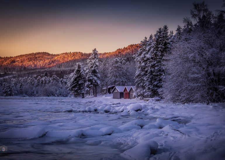 Gefrorenes Ende eines Fjords im Winterlicht. Vereiste Wasserfläche, stille Bootshäuser und ruhige Winterlandschaft bei klarem Licht.