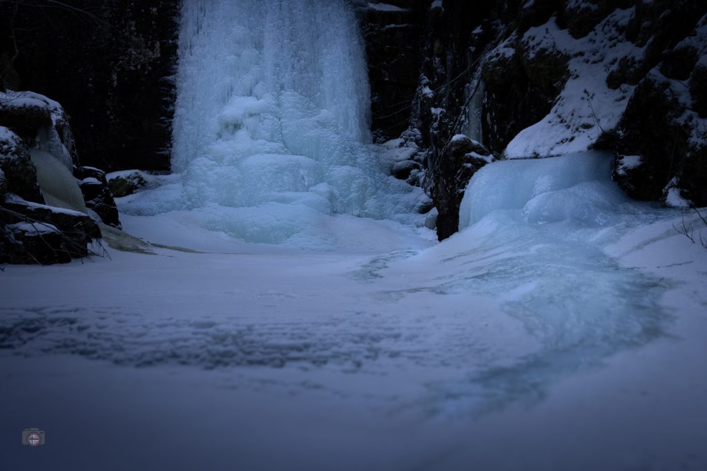 Ein gefrorener Wasserfall in einer schattigen Schlucht. Das Eis hat das fallende Wasser in eine massive, blaue Struktur verwandelt. Schnee bedeckt den Boden, dunkle Felsen rahmen die Szene. Die Atmosphäre wirkt still und zeitlos – als würde der Winter den Atem anhalten.