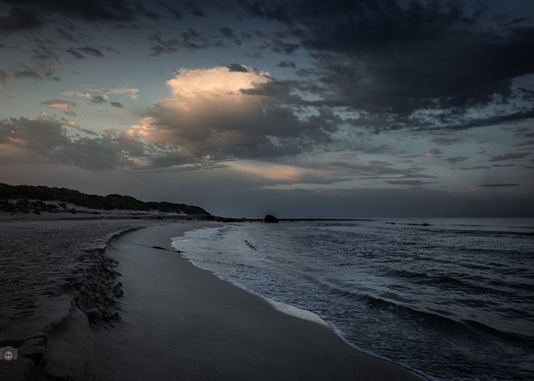 Langzeitbelichtung eines menschenleeren Strandes mit moosbewachsenen Steinen und einem großen Felsen am frühen Morgen