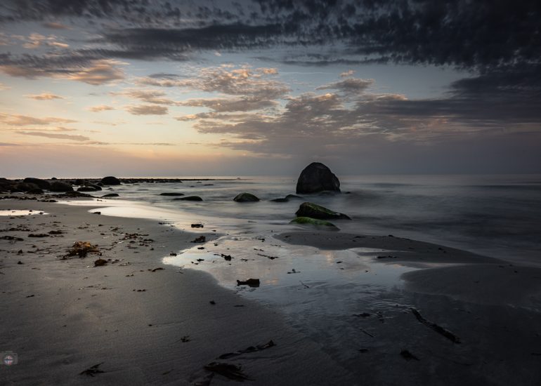 Langzeitbelichtung eines menschenleeren Strandes mit moosbewachsenen Steinen und einem großen Felsen am frühen Morgen
