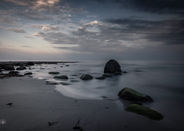 Langzeitbelichtung eines menschenleeren Strandes mit moosbewachsenen Steinen und einem großen Felsen am frühen Morgen