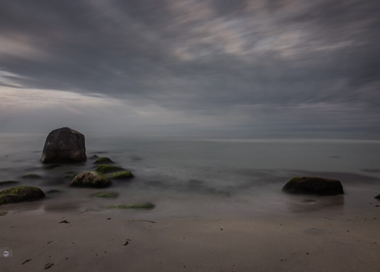 Langzeitbelichtung eines menschenleeren Strandes mit moosbewachsenen Steinen und einem großen Felsen am frühen Morgen