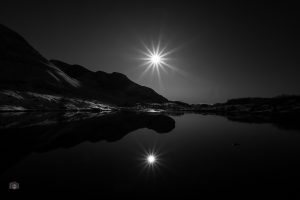 Sonne über einer Berglandschaft im Gegenlicht mit Spiegelung im ruhigen Wasser eines Bergsees.