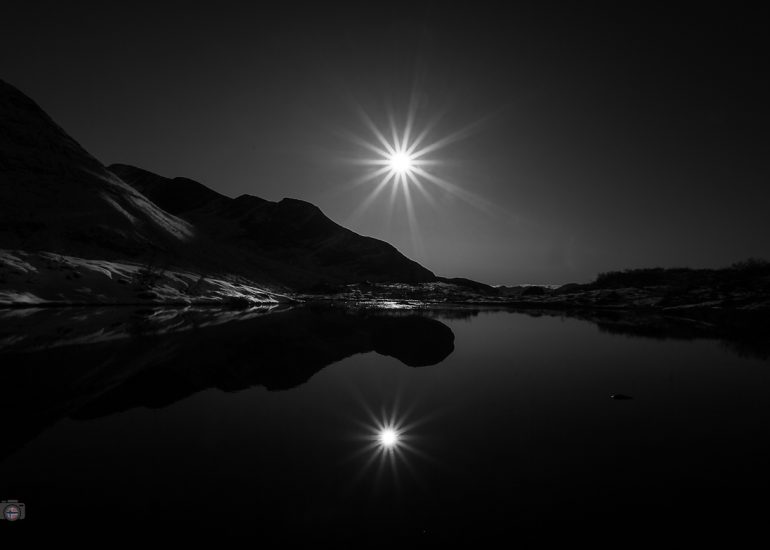 Sonne über einer Berglandschaft im Gegenlicht mit Spiegelung im ruhigen Wasser eines Bergsees.