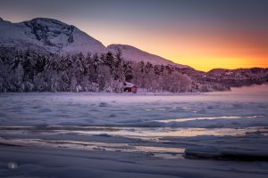 Winterlandschaft mit gefrorenem Fjord bei Sonnenaufgang. Warmes Licht am Horizont, schneebedeckte Berge und erste Anzeichen des nahenden Frühlings.