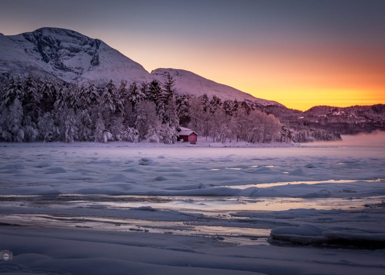 Winterlandschaft mit gefrorenem Fjord bei Sonnenaufgang. Warmes Licht am Horizont, schneebedeckte Berge und erste Anzeichen des nahenden Frühlings.