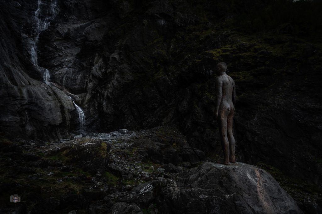 Skulptur am Felsen neben einem Wasserfall in dunkler Stimmung mit sichtbarer Wasserbewegung
