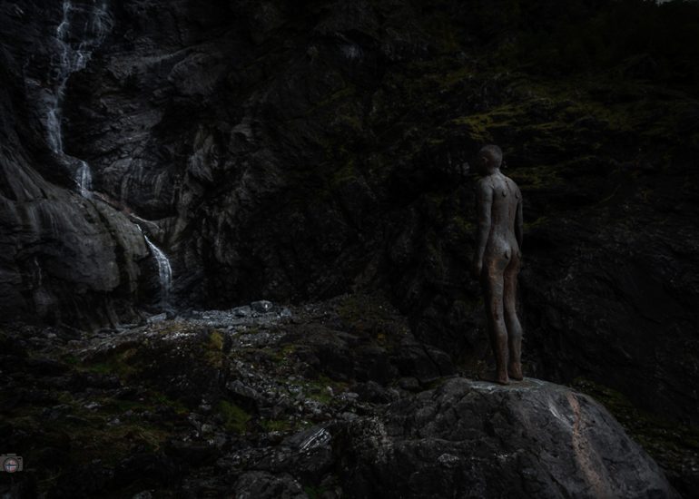 Skulptur am Felsen neben einem Wasserfall in dunkler Stimmung mit sichtbarer Wasserbewegung