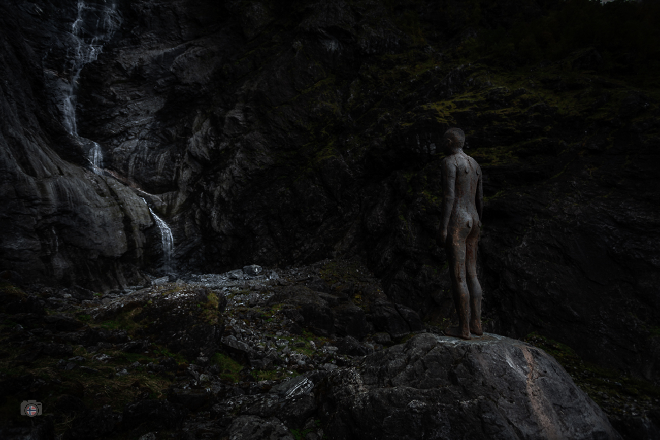 Skulptur am Felsen neben einem Wasserfall in dunkler Stimmung mit sichtbarer Wasserbewegung