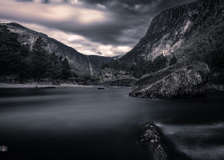 Langzeitbelichtung eines Flusses mit ruhigem Wasser, dunklen Bergen und Felsen im Vordergrund
