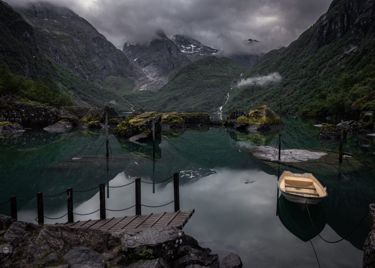 Stiller Bergsee mit spiegelnden Felsen und senkrechten Pfählen vor bewaldeten, wolkenverhangenen Bergen.