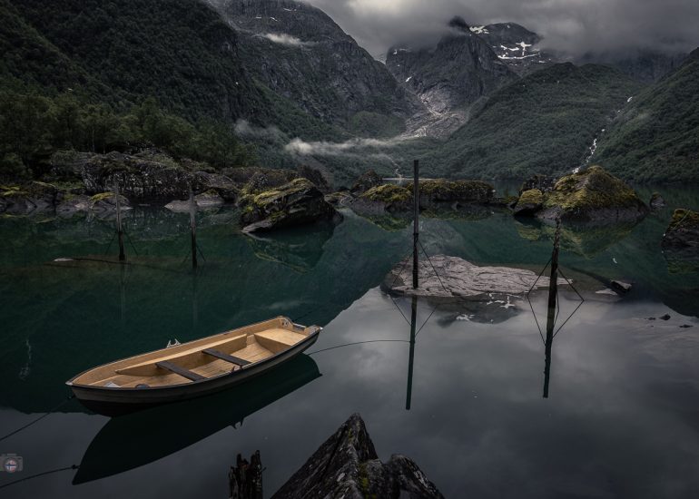 Stiller Bergsee mit spiegelnden Felsen und senkrechten Pfählen vor bewaldeten, wolkenverhangenen Bergen.