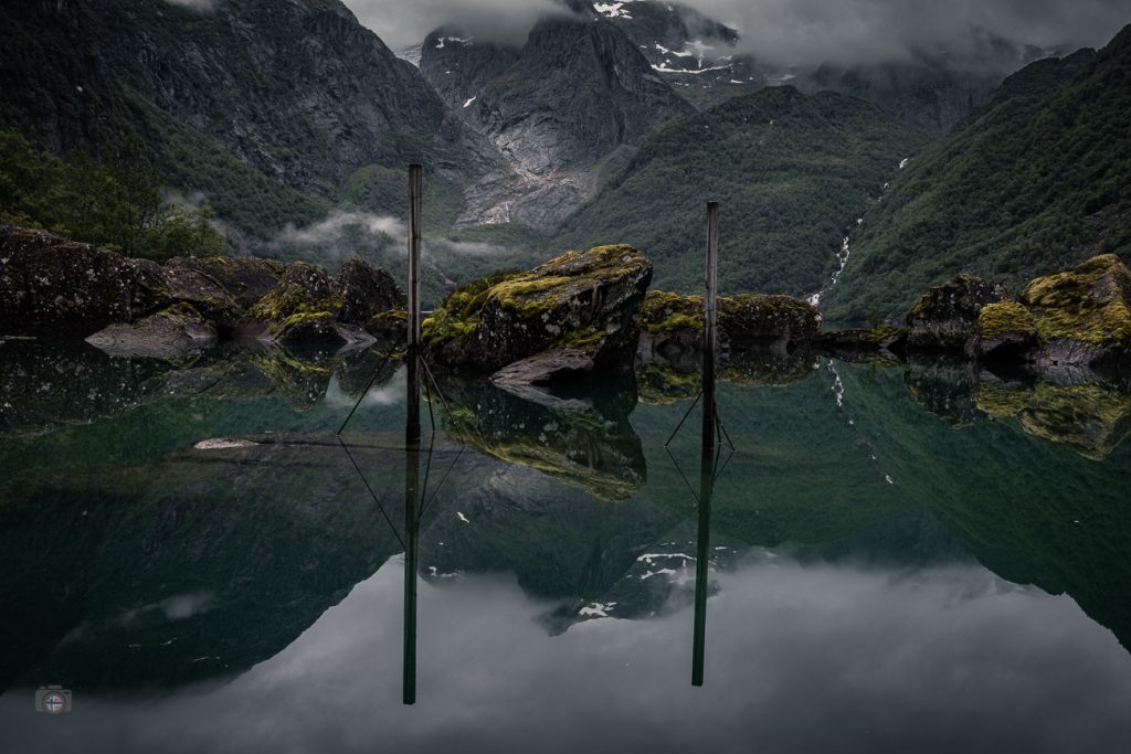 Stiller Bergsee mit spiegelnden Felsen und senkrechten Pfählen vor bewaldeten, wolkenverhangenen Bergen.