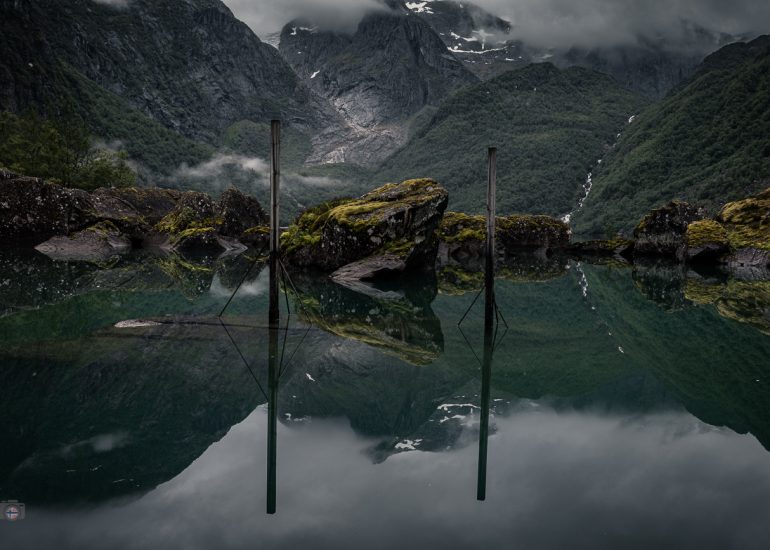 Dunkler Bergsee mit moosbewachsenen Felsen, Pfählen und spiegelnden Bergen unter tief hängenden Wolken