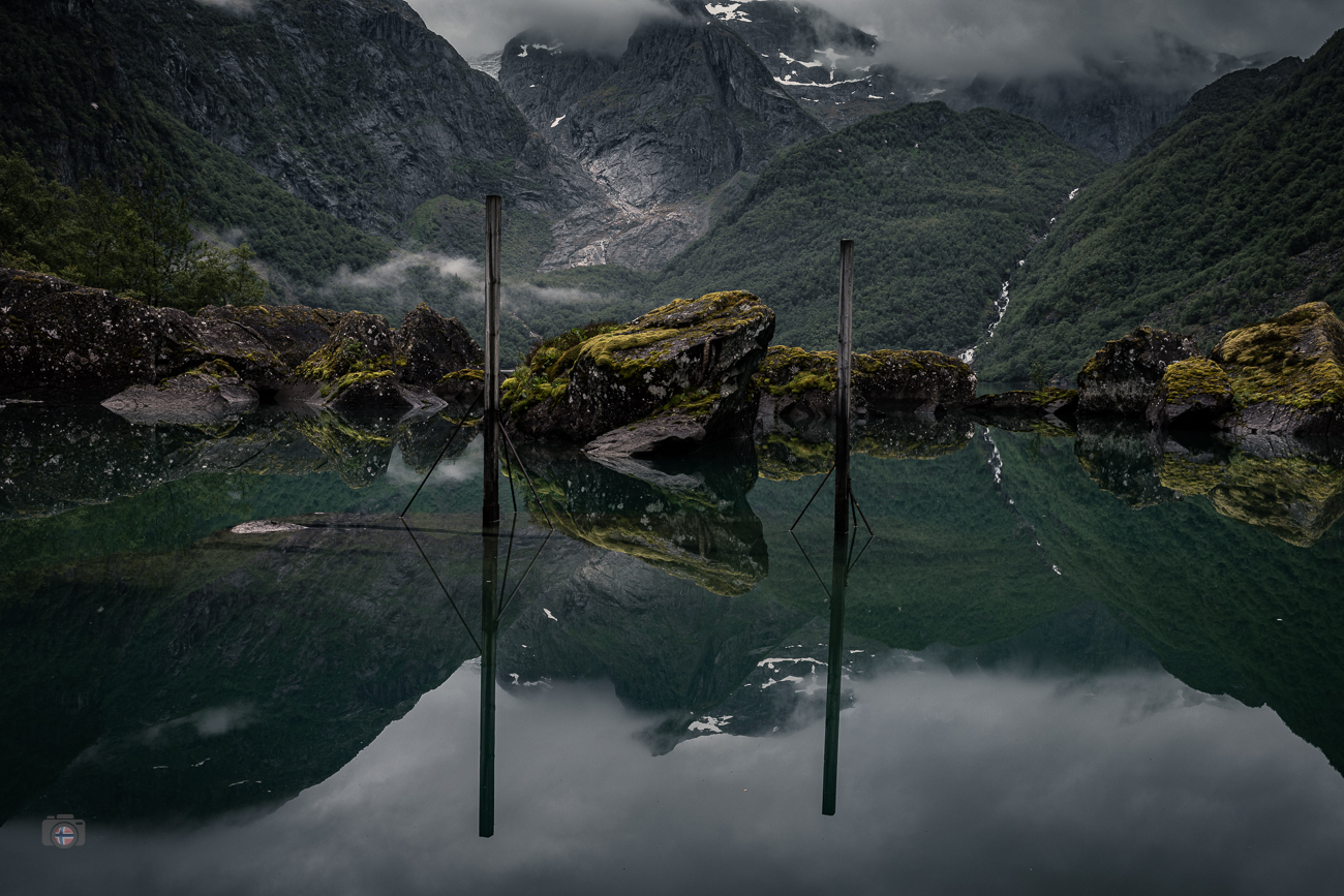 Dunkler Bergsee mit moosbewachsenen Felsen, Pfählen und spiegelnden Bergen unter tief hängenden Wolken
