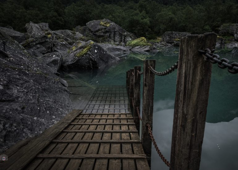 Stiller Bergsee mit spiegelnden Felsen und senkrechten Pfählen vor bewaldeten, wolkenverhangenen Bergen.
