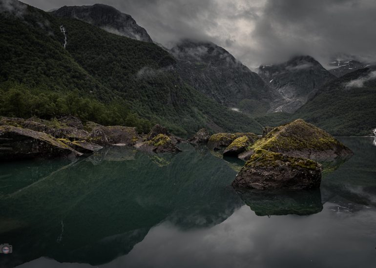 Stiller Bergsee mit spiegelnden Felsen und senkrechten Pfählen vor bewaldeten, wolkenverhangenen Bergen.