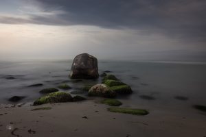 Langzeitbelichtung eines menschenleeren Strandes mit moosbewachsenen Steinen und einem großen Felsen am frühen Morgen
