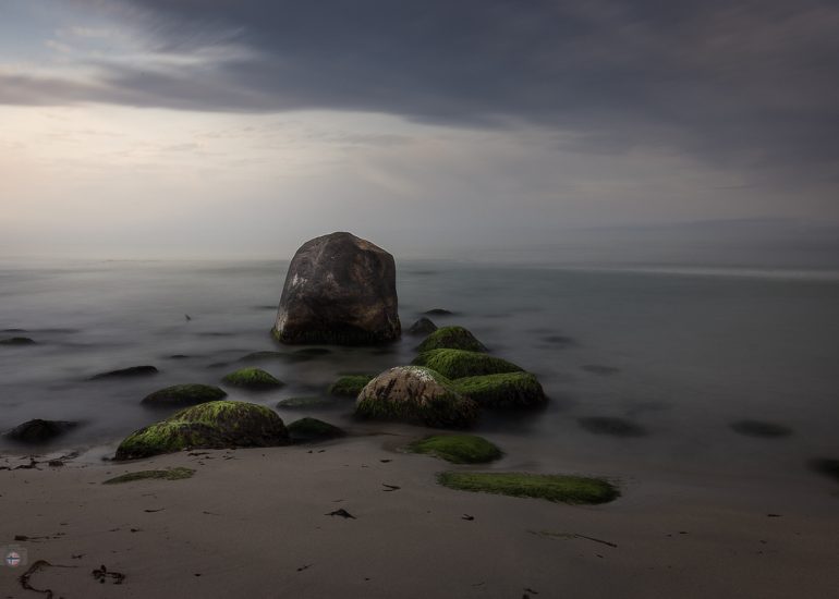 Langzeitbelichtung eines menschenleeren Strandes mit moosbewachsenen Steinen und einem großen Felsen am frühen Morgen
