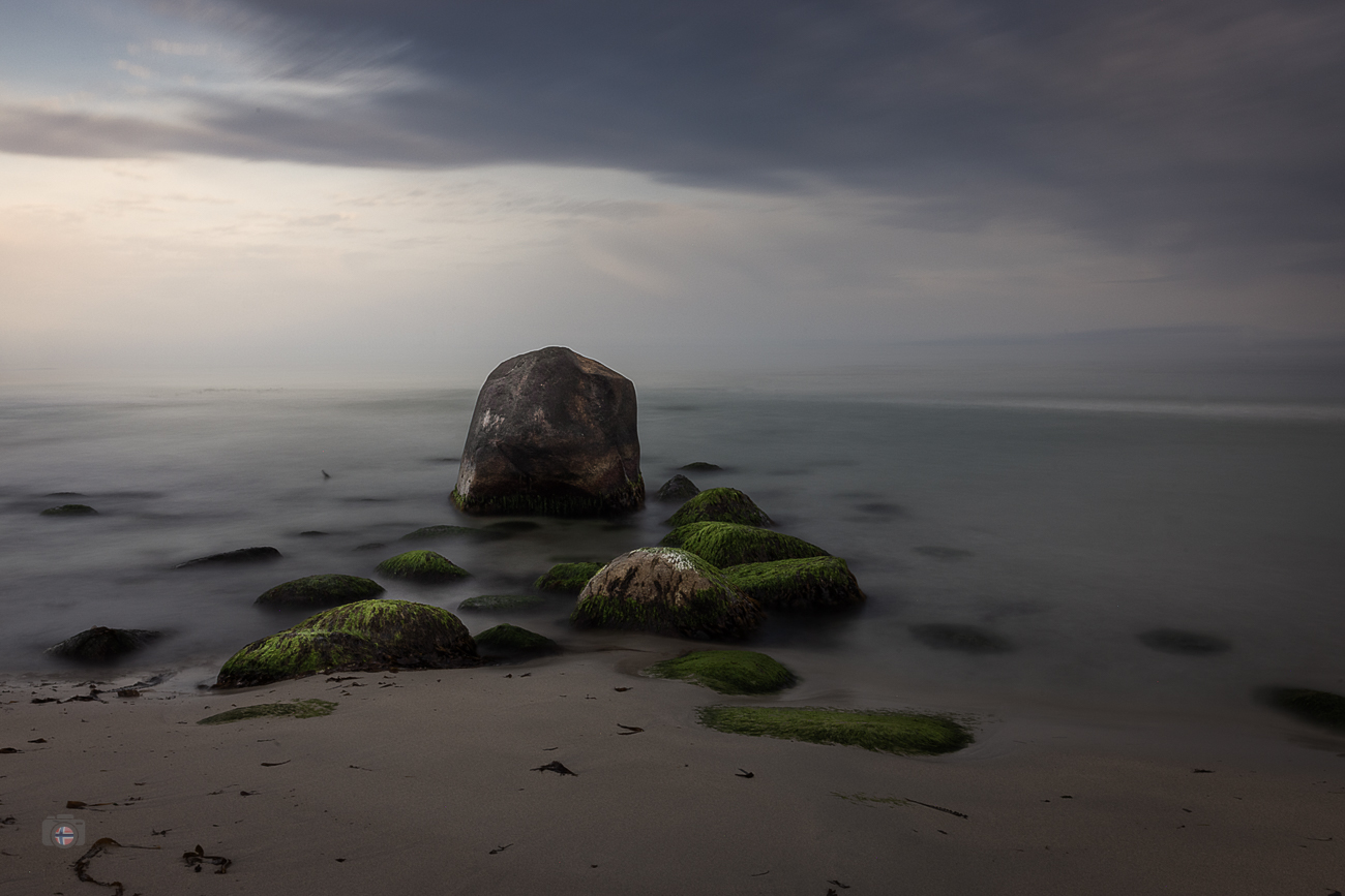 Langzeitbelichtung eines menschenleeren Strandes mit moosbewachsenen Steinen und einem großen Felsen am frühen Morgen