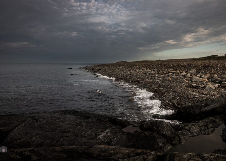 Tiefe Perspektive über unzählige runde Steine eines Küstenstrandes mit Meer und bewölktem Himmel im Hintergrund.