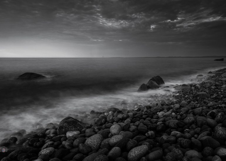 Tiefe Perspektive über unzählige runde Steine eines Küstenstrandes mit Meer und bewölktem Himmel im Hintergrund.