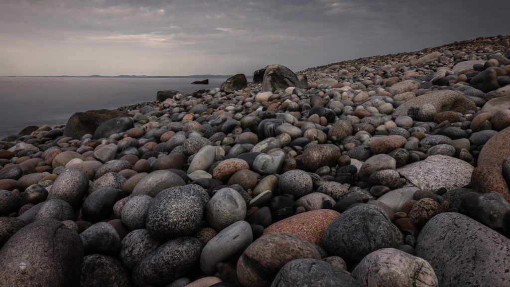 Tiefe Perspektive über unzählige runde Steine eines Küstenstrandes mit Meer und bewölktem Himmel im Hintergrund.