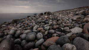 Tiefe Perspektive über unzählige runde Steine eines Küstenstrandes mit Meer und bewölktem Himmel im Hintergrund.