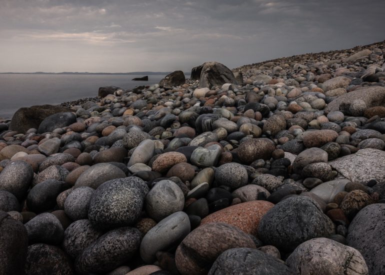 Tiefe Perspektive über unzählige runde Steine eines Küstenstrandes mit Meer und bewölktem Himmel im Hintergrund.