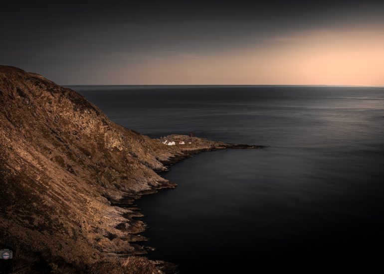 Dunkle norwegische Küstenlandschaft mit kleiner Häusergruppe am Meer und warmem Licht am Horizont.