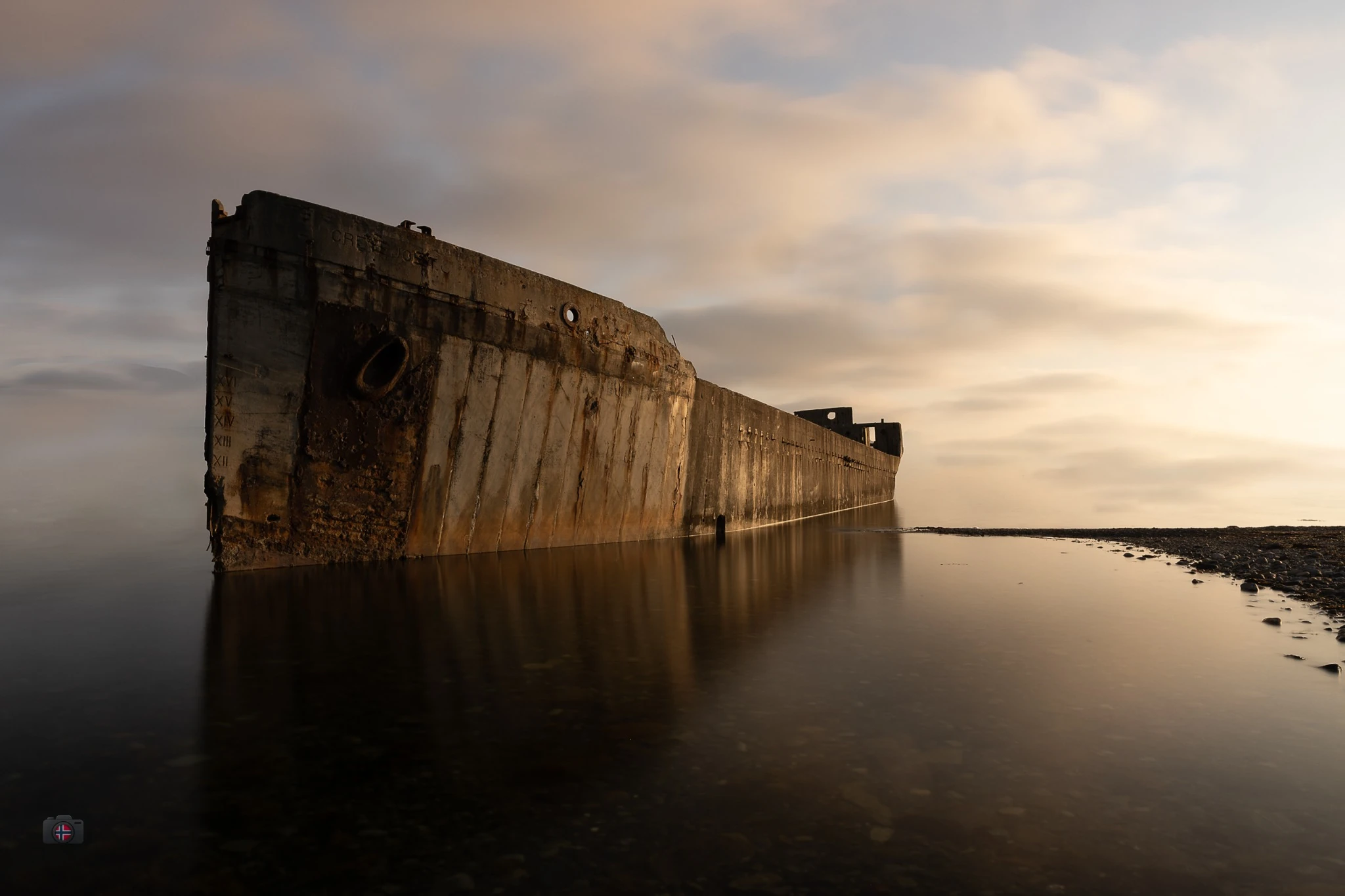 Verrosteter Rumpf eines Schiffswracks im ruhigen Meer bei warmem Abendlicht und weich verwischtem Himmel.