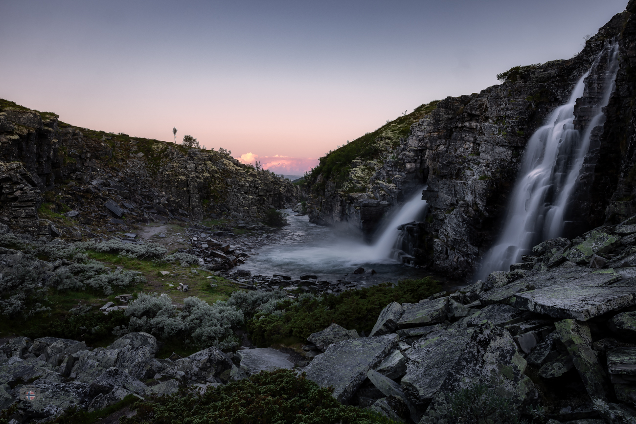 Zwischen den dunklen Felsen rauscht das Wasser durch die noch kühle Landschaft. Erst weit hinten am Horizont erscheint ein Hauch von Farbe. 
