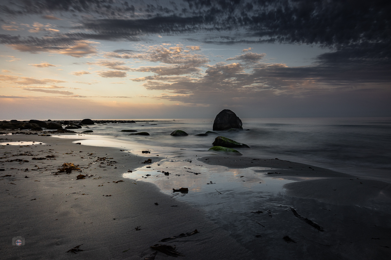 Felsen an einem ruhigen Sandstrand im frühen Morgenlicht mit weich gezeichneter Brandung und Wolken am Himmel.