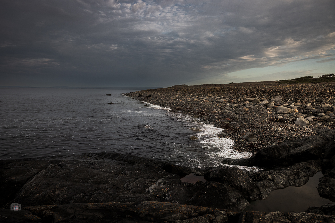 Unzählige Steine formen eine Küste, die sich langsam im Meer verliert. Unter dem schweren Himmel wirkt die Landschaft still und unscheinbar
