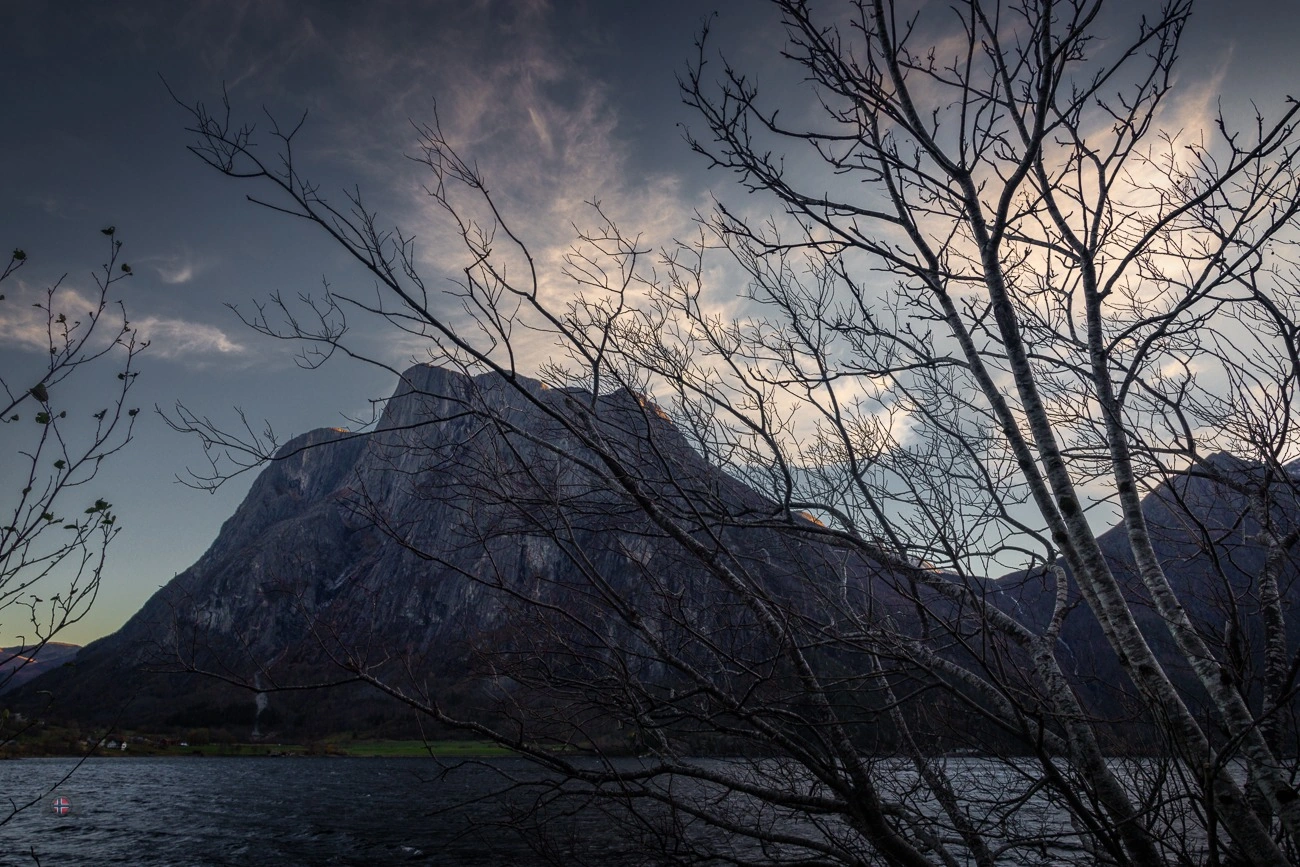 Blick durch kahle Zweige auf steile Berge und einen Bergsee im herbstlichen Licht