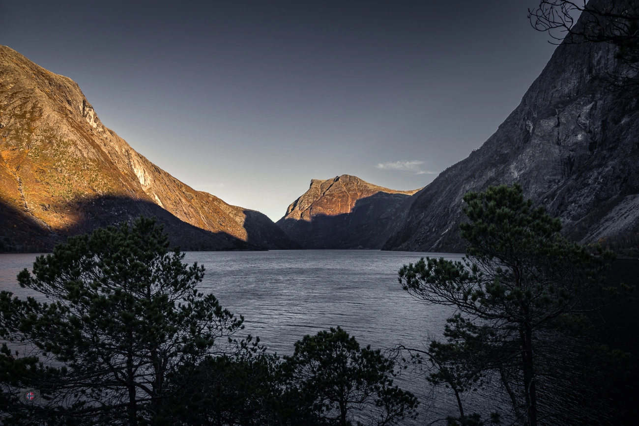 Goldenes Licht auf dem Bergkamm über einem dunklen Bergsee, mit Steinen am Ufer