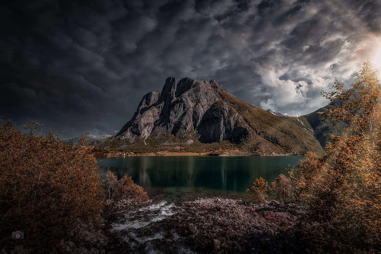 Herbstliche Berglandschaft in Norwegen mit goldbraunen Bäumen am See und Wolken über den Gipfeln.
