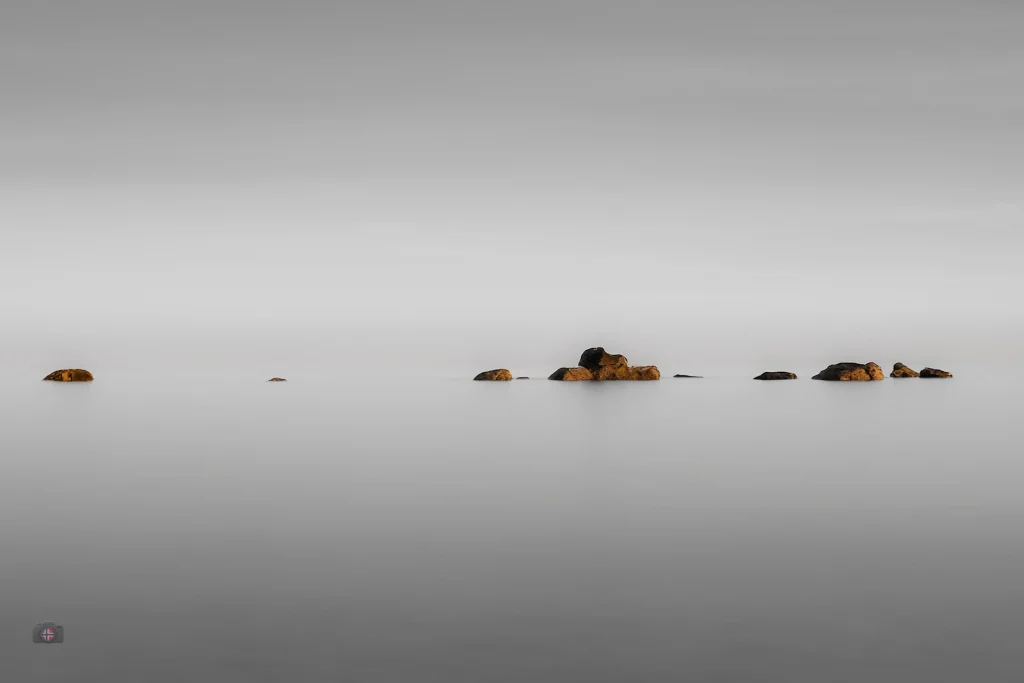 Kleine Felsen als feine Linie in glatter, heller Meeresfläche mit viel negativem Raum.